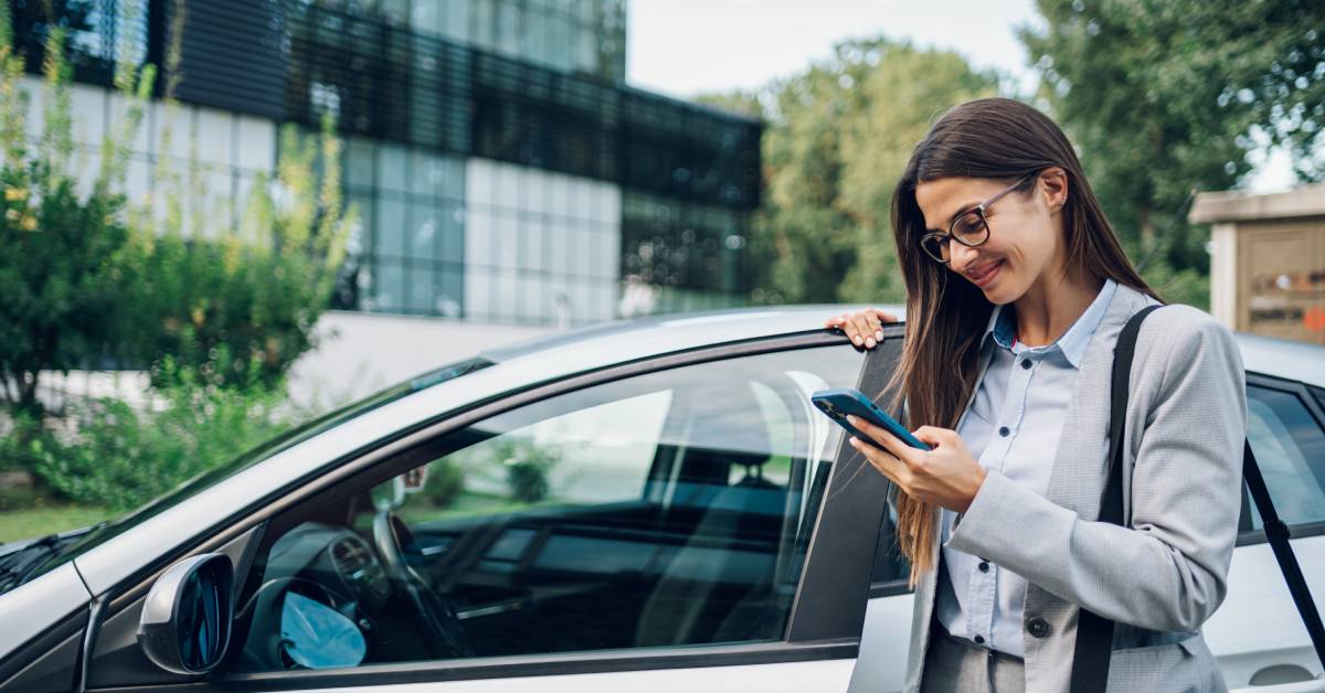 A business woman in a suit holding her smartphone as she gets into her car. She is parked outside an office building.